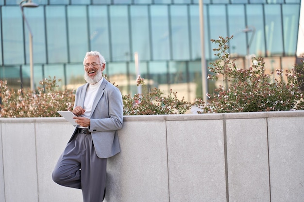 Happy old business man holding digital tablet standing outside city buildings