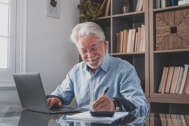 행복한 노년의 백인 사업가가 노트북으로 온라인 웨비나 팟캐스트를 시청하고 교육 과정 회의 통화를 하며 업무용 책상에 메모를 하고 전자 학습 개념을 보며 웃고 있습니다.
