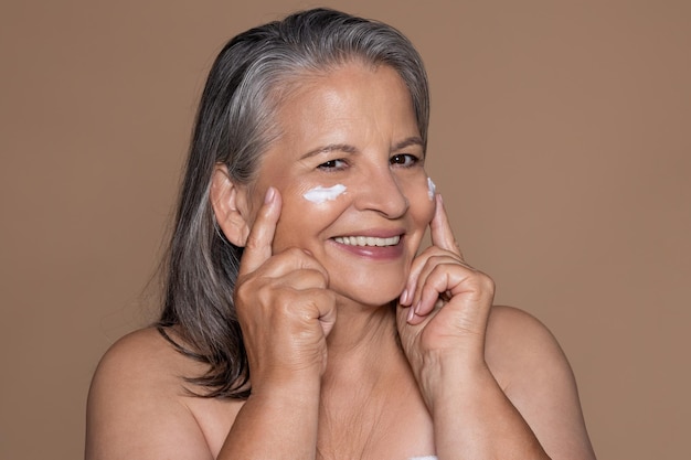 Happy old caucasian woman with gray hair enjoys antiaging treatment moisturizing cosmetics
