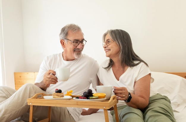 Happy old european husband and wife have breakfast enjoy coffee cups sit on bed