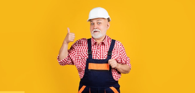 Счастливый старик строитель в шлеме на желтом фоне большой палец вверх