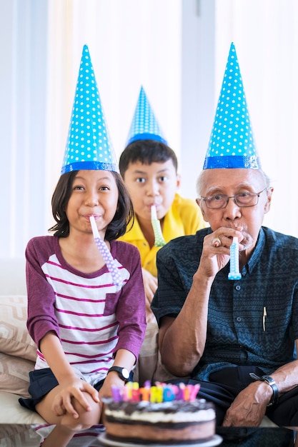 Happy old man celebrating his birthday with his grandchildren at home while wearing birthday hat and blowing trumpet