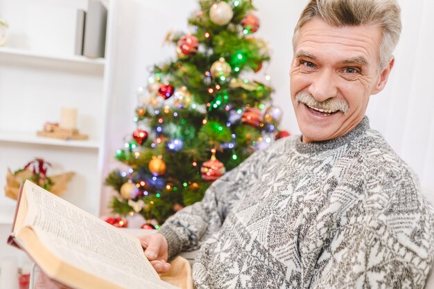 The happy old man hold a book near the christmas tree