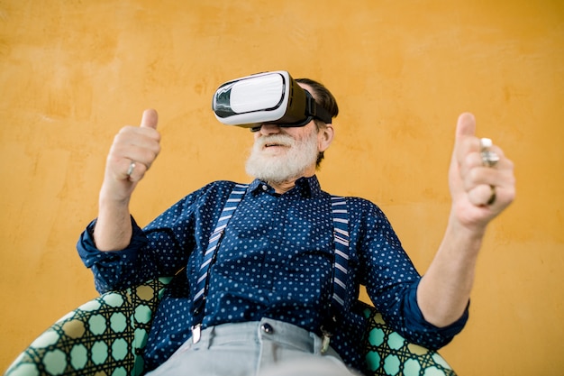 happy old man with well-groomed beard showing his thumbs up while using virtual reality goggles for watching film. Studio shot on yellow background. VR technologies