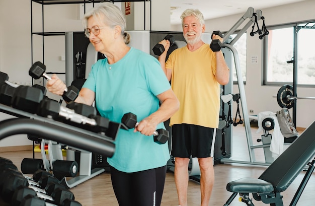 Happy old senior couple in sportswear exercising with dumbbells in gym doing exercises to stay fit