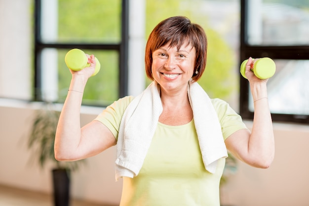 Happy older woman in sports wear training with dumbbells indoors on the window background