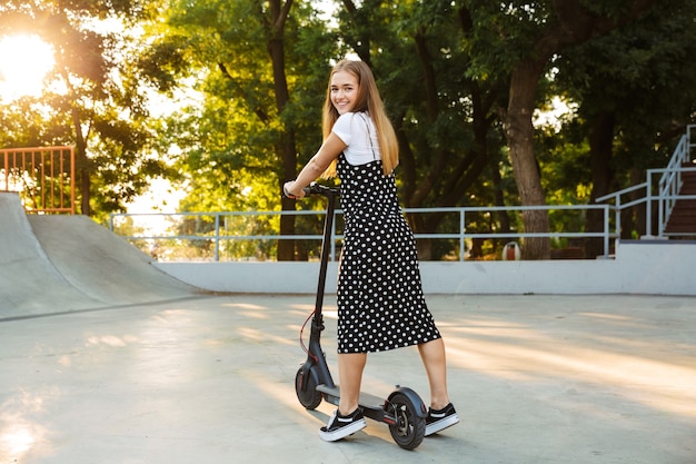 happy optimistic cheerful teenage girl in park walking on scooter.