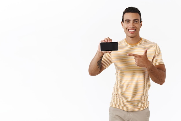 Happy, outgoing handsome athletic man in t-shirt, holding smartphone horizontally and pointing finger at display