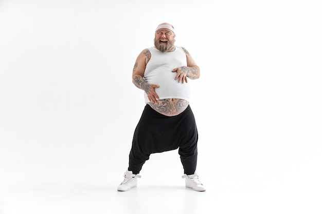 Photo happy overweight man with big belly and tattoos in sports wear is holding his stomach