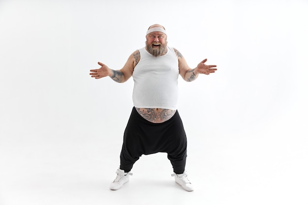 Happy overweight man with big belly and tattoos in sports wear posing on white background