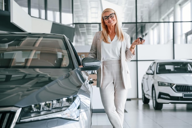 キーを手にした新しい自動車の幸せな所有者白いフォーマルな服を着た女性が自動車販売店にいる