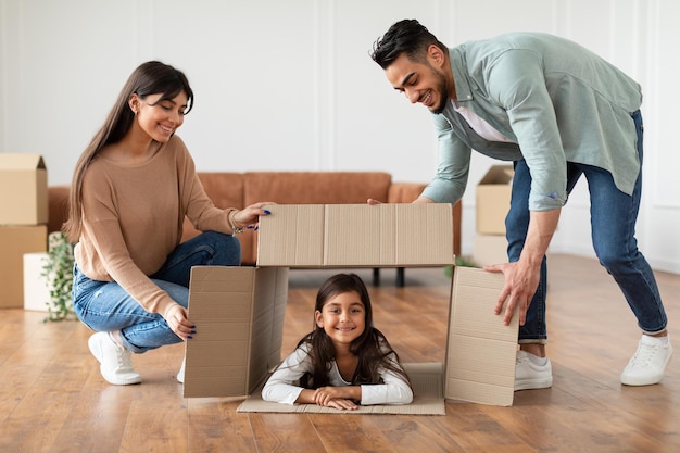 Happy parents playing with cute little kid on moving day, laughing spouses having fun with daughter sitting in carton box in living room, family tenants renters imitating house. Relocating new home