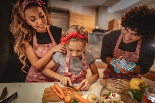 Happy parents and their daughter cooking together in the kitchen while little girl trying to cut carrot.