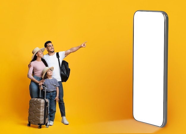 Happy parents with little daughter carrying suitcases pointing at phone