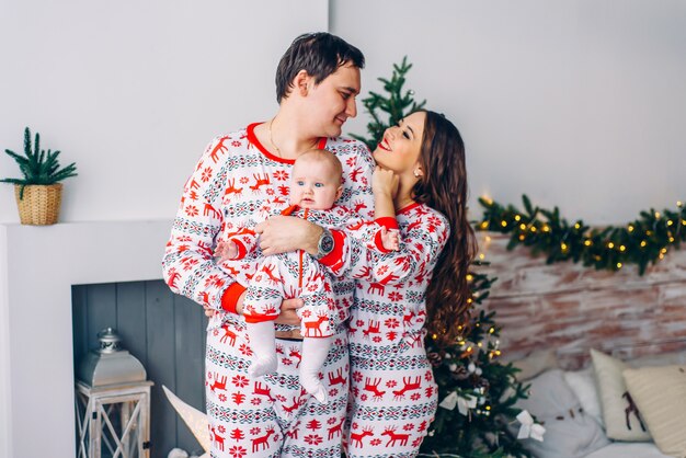 Happy parents with their little daughter in holiday clothing with printed deers and snowflakes in cozy room with a Christmas tree