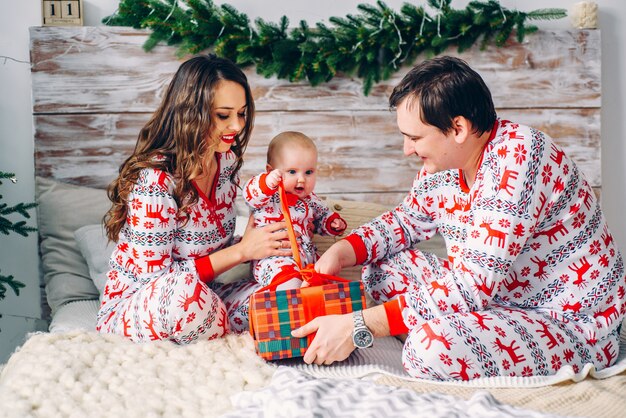 Genitori felici con la loro piccola figlia in vestiti di festa con cervi e fiocchi di neve stampati che disimballano il regalo di natale nella stanza accogliente