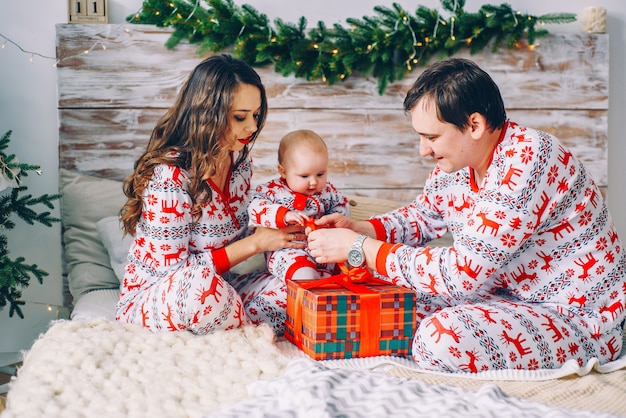 Happy parents with their little daughter in holiday clothing with printed deers and snowflakes unpacking Christmas gift in cozy room 