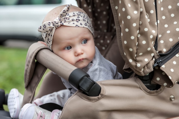 Happy pensive emotional eight-month-old blue-eyed girl sit in stroller on walk and wait for mom