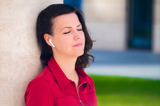 Happy pensive girl, young beautiful woman relaxing listening to music in wireless new modern earphones with closed eyes, enjoy song, good weather, dream outdoors. Calm, relaxation, technology concept