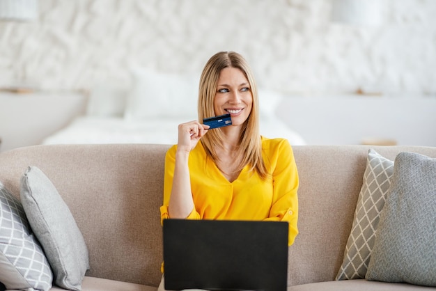 Happy pensive millennial european blond lady in yellow clothes with laptop thinks about buying in