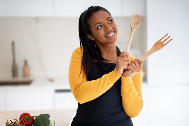 野菜とテーブルの近くのエプロンで幸せな物思いにふける若いアフリカ系アメリカ人女性クックは、木製のフォークで夢を考えています