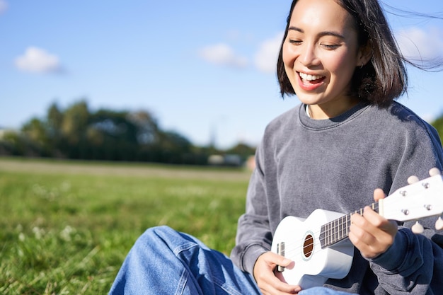 Happy people and hobbies smiling asian girl playing ukulele guitar and singing sitting in park outdo