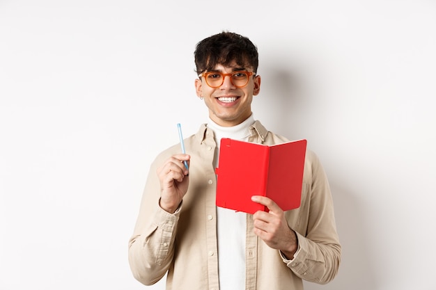 Happy person in glasses writing in journal, holding pen and diary, smiling, plan a schedule, standing with planner on white wall.