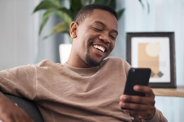 Happy phone and man in a living room texting laughing and reading social media post in his home Day off black man and online chat on smartphone text and funny message on dating website or app