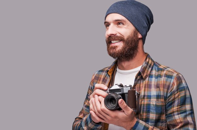 Happy photographer. Handsome young bearded man holding old-fashioned camera and looking away with smile while standing against grey background