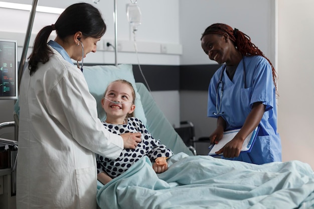 Photo happy physician expert and nurse consulting ill little girl health condition while smiling together inside hospital pediatric ward. multiethnic clinic staff examining sick kid resting in patient bed.