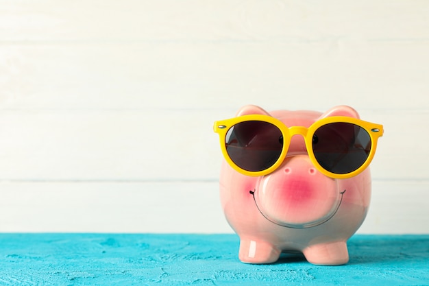 Happy piggy bank with sunglasses on color table against white wooden background