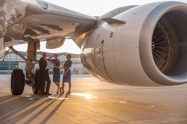 Happy pilot and two pretty stewardesses standing together, leaning on an airplane and smiling at camera after landing or before departure. Aircraft, aircrew, occupation concept