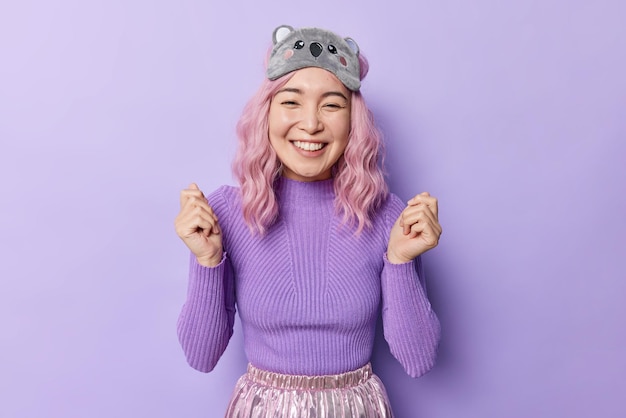 Happy pink haired young Asian woman shakes arms smiles gladfully being in good mood after healthy sleep wears blidfold and casual jumper isolated over purple background Positive human emotions