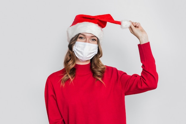 Happy playful young woman in santa hat, red sweater and medical protective respiratory face mask holding and pulling balabon isolated on gray studio background