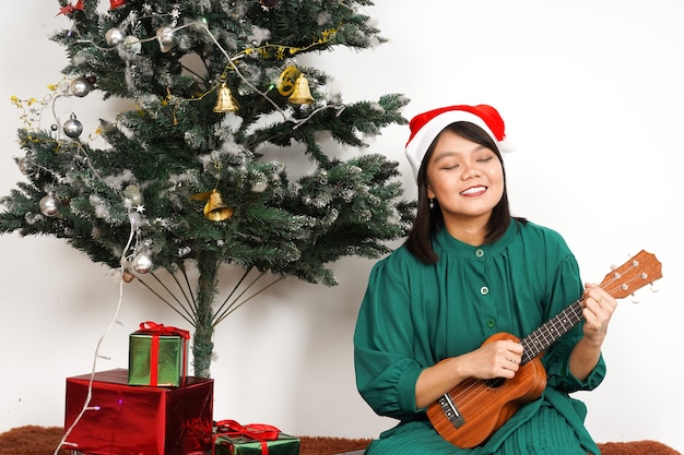 Счастливый и играет на гавайской гитаре Красивая азиатская женщина в зеленом платье и шляпе Санта-Клауса
