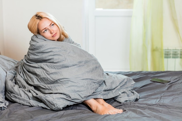 写真 快適なベッドで暖かい柔らかい灰色の毛布で覆われて座っている幸せな幸せな女性。寝室で時間を過ごす美しい眠そうな女性。休息、リラックス、週末、怠惰の概念。