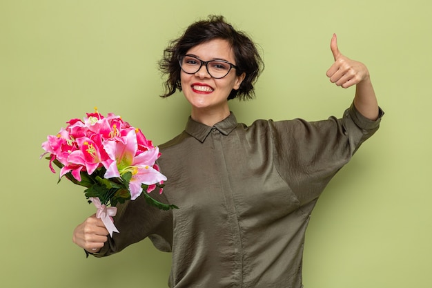 Happy and pleased woman with short hair holding bouquet of flowers looking at camera smiling cheerfully showing thumbs up celebrating international women's day march 8 standing over green background