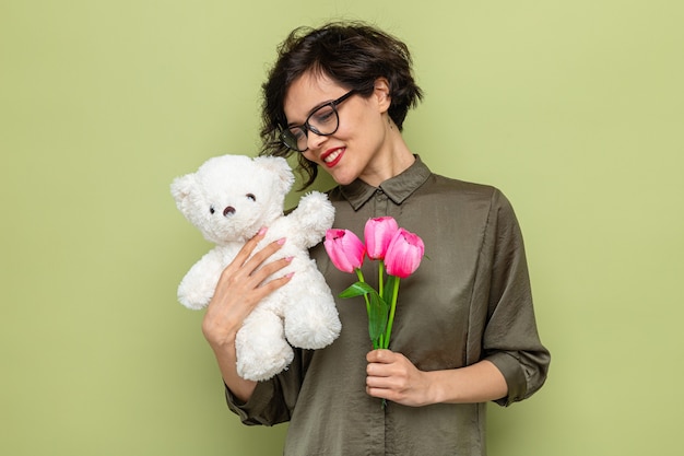 Happy and pleased woman with short hair holding bouquet of tulips and teddy bear smiling cheerfully celebrating international women's day march 8 standing over green background