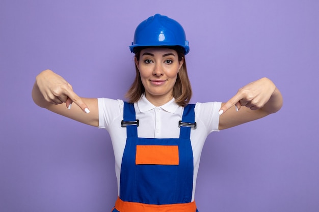 Felice e compiaciuta giovane donna costruttore in uniforme da costruzione e casco di sicurezza che sembra sorridente fiduciosa che indica se stessa