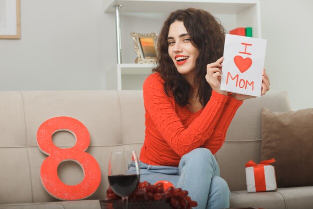 Felice e felice giovane donna in abiti casual seduto su un divano con il numero otto e presente che tiene biglietto di auguri sorridente allegramente celebrando la giornata internazionale della donna 8 marzo