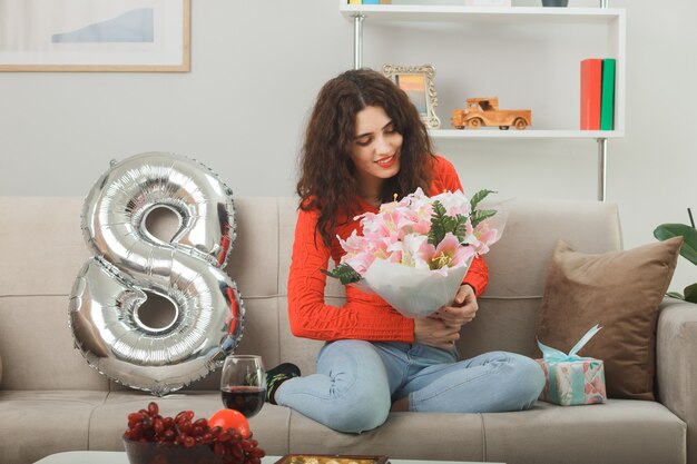 Happy and pleased young woman in casual clothes smiling\
cheerfully sitting on a couch with number eight shaped balloon\
holding bouquet of flowers celebrating international womens day\
march 8