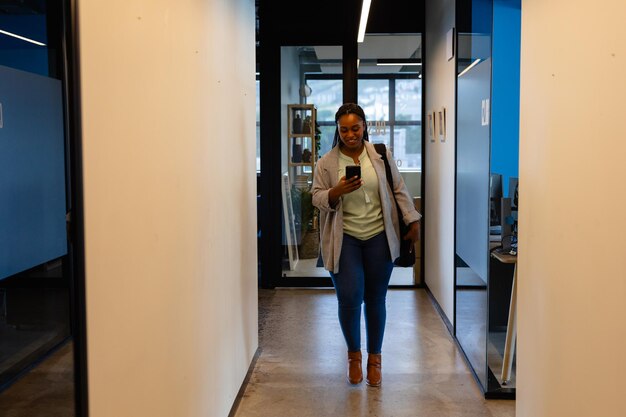 Фото Счастливая афроамериканская деловая женщина с смартфоном в коридоре офиса. офис, общение, бизнес, профессиональность и работа, неизменны.