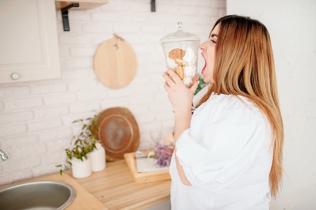 Happy plus size model in jeans and a white T-shirt, holding a sweetness in her hands, opening her mouth in a stylish bright kitchen. Young plump woman in casual outfit. The body is positive. The conce