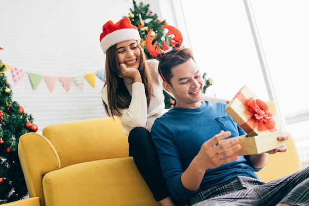 아시아 여성의 행복한 초상화는 그의 남자친구를 크리스마스 선물로 놀라게합니다. 아침에 집에서 크리스마스 트리와 함께 복사 공간을 가진 배경 이미지로 거실에서 미소 짓고 있습니다.