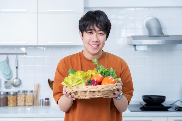 Счастливый портрет азиатского молодого человека с корзиной овощей стоящего веселого приготовления пищи и наслаждаться приготовлением пищи с овощами, стоя на кухне в кондоминиуме или дома