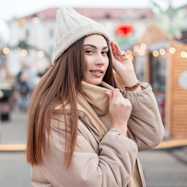 Il ritratto felice di una bella ragazza con un cappello lavorato a maglia e una giacca invernale beige con una sciarpa cammina per la città