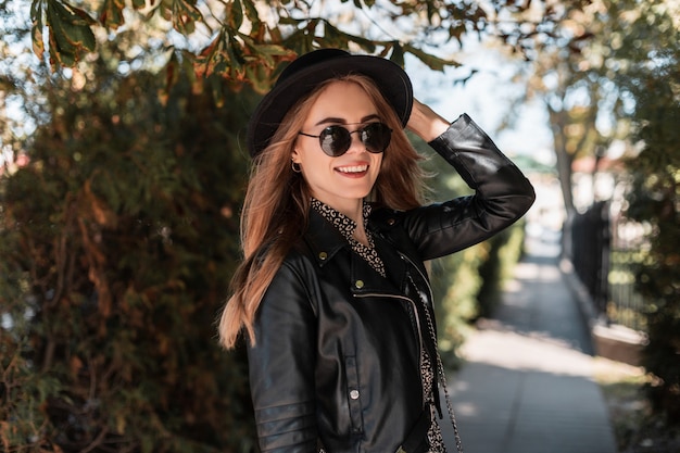 Happy portrait of a beautiful young girl model in fashionable clothes look with a leather jacket and stylish sunglasses walks in the autumn park