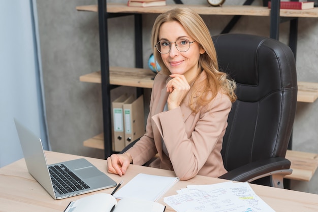 Ritratto felice di una giovane donna di affari sorridente che si siede sulla sedia nel luogo di lavoro con il computer portatile e le carte sul tavolo