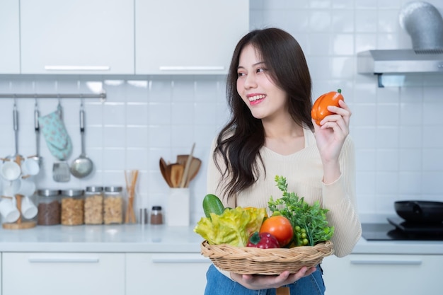 Счастливый портрет молодой азиатской женщины, держащей корзину с овощами, стоящей, весело готовя еду и наслаждаясь приготовлением еды с овощими, стоя на кухне, в кондоминиуме или дома.