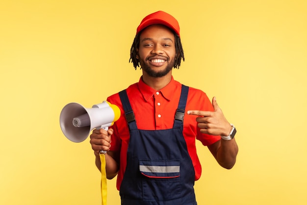 Happy positive bearded workman wearing blue overalls and red T-shirt looking at camera, holding megaphone in hands, announcing advertisement. Indoor studio shot isolated on yellow background.